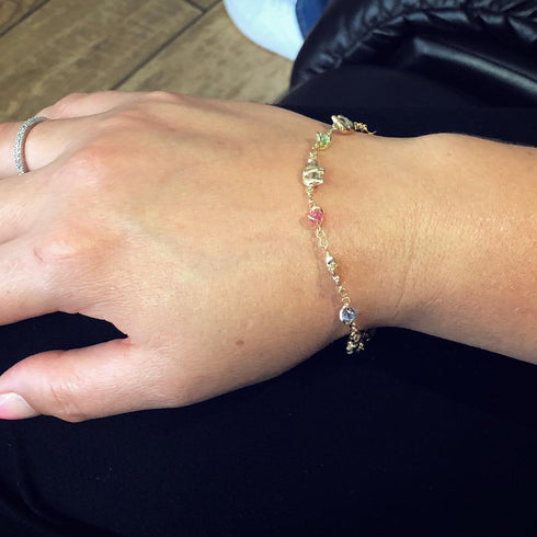 A hand adorned with a delicate Good Luck Elephant Bracelet with Multicolor Birthstones rests on dark fabric. A ring graces the middle finger, while the background reveals a wooden floor and part of an armchair.